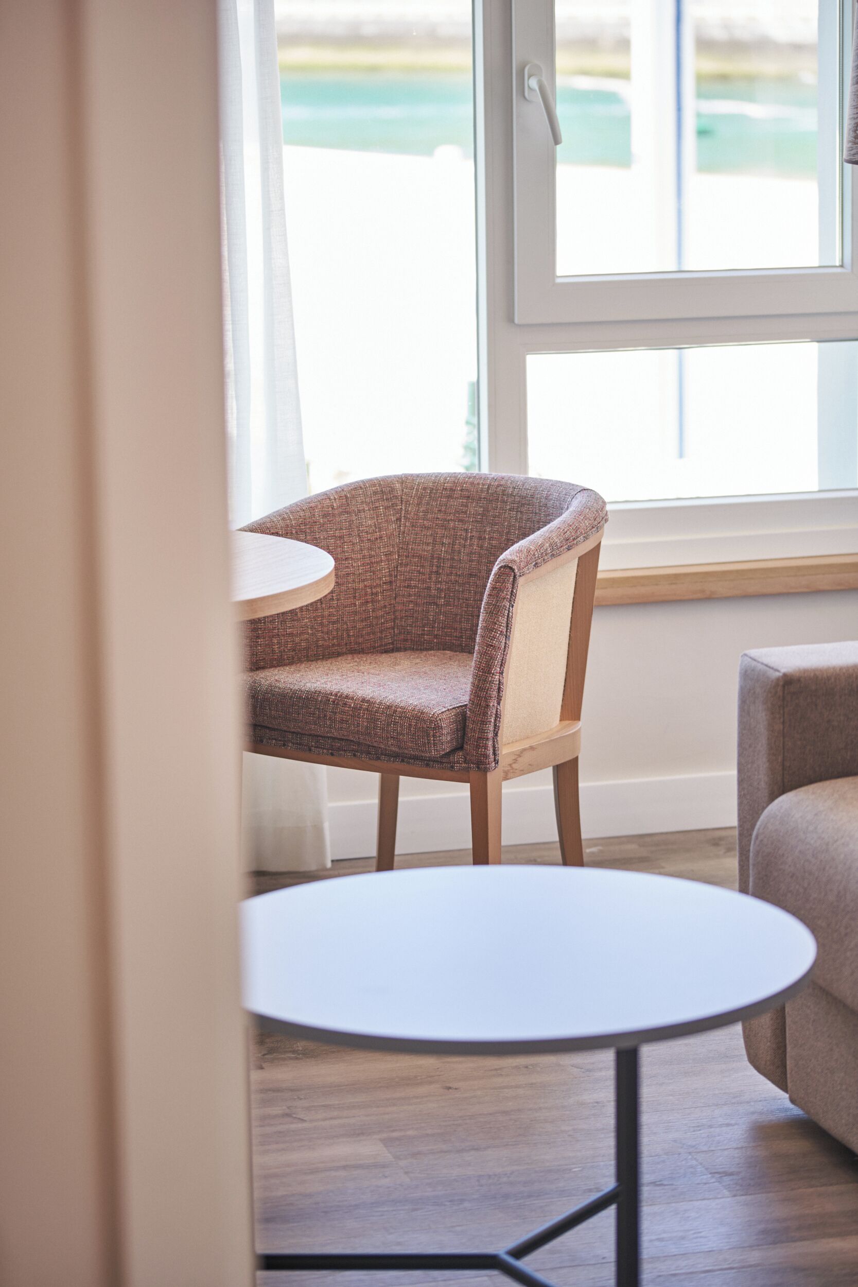 Een kamer met een stoel en een kleine ronde tafel, een raam met uitzicht en een houten vloer.