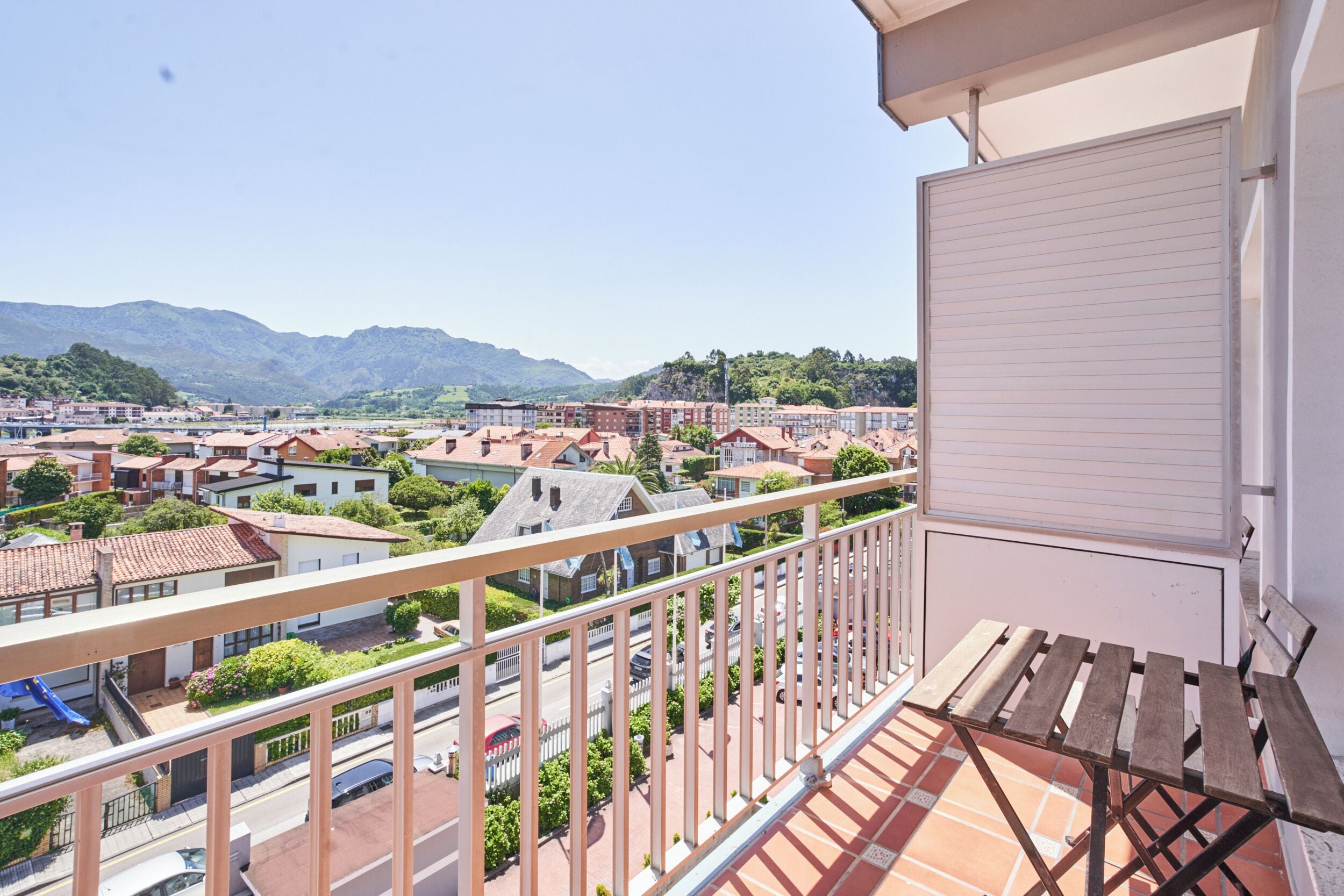 A balcony with a view of a residential area, a closed shutter, and a wooden bench.