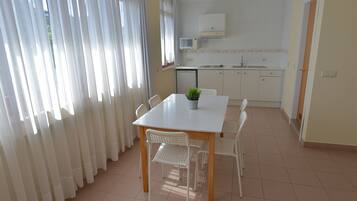 A compact kitchen with white cabinets and a microwave, a dining area with a white table and chairs, and a window with white curtains.