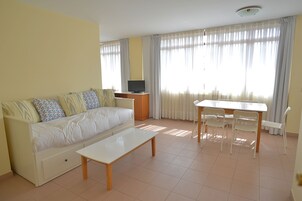 Uma sala de estar com sofá, televisão em móvel, mesa de jantar com cadeiras e grandes janelas com cortinas.