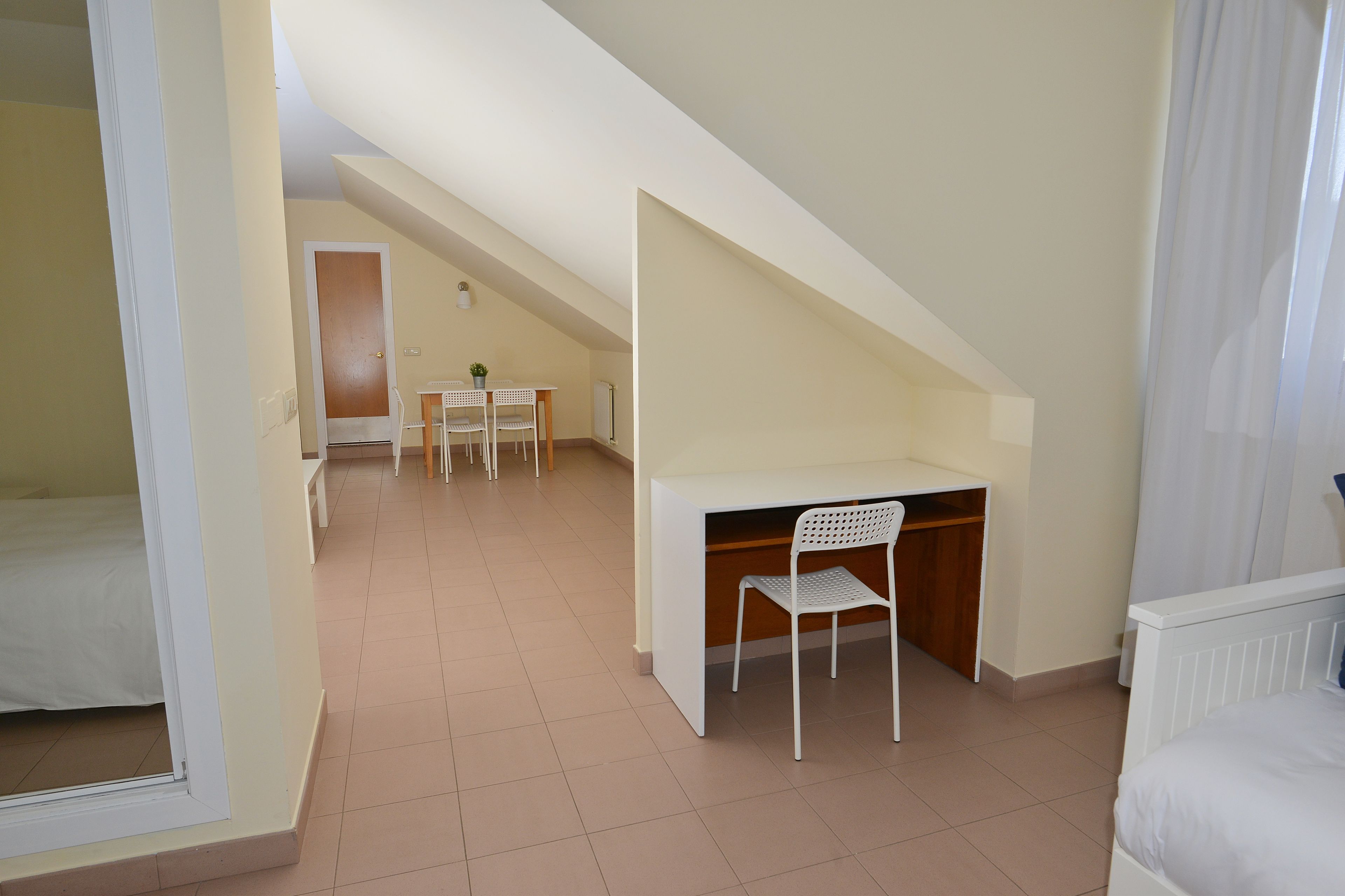 A bedroom with a bed, a desk with a chair, and a small table with chairs in the background.