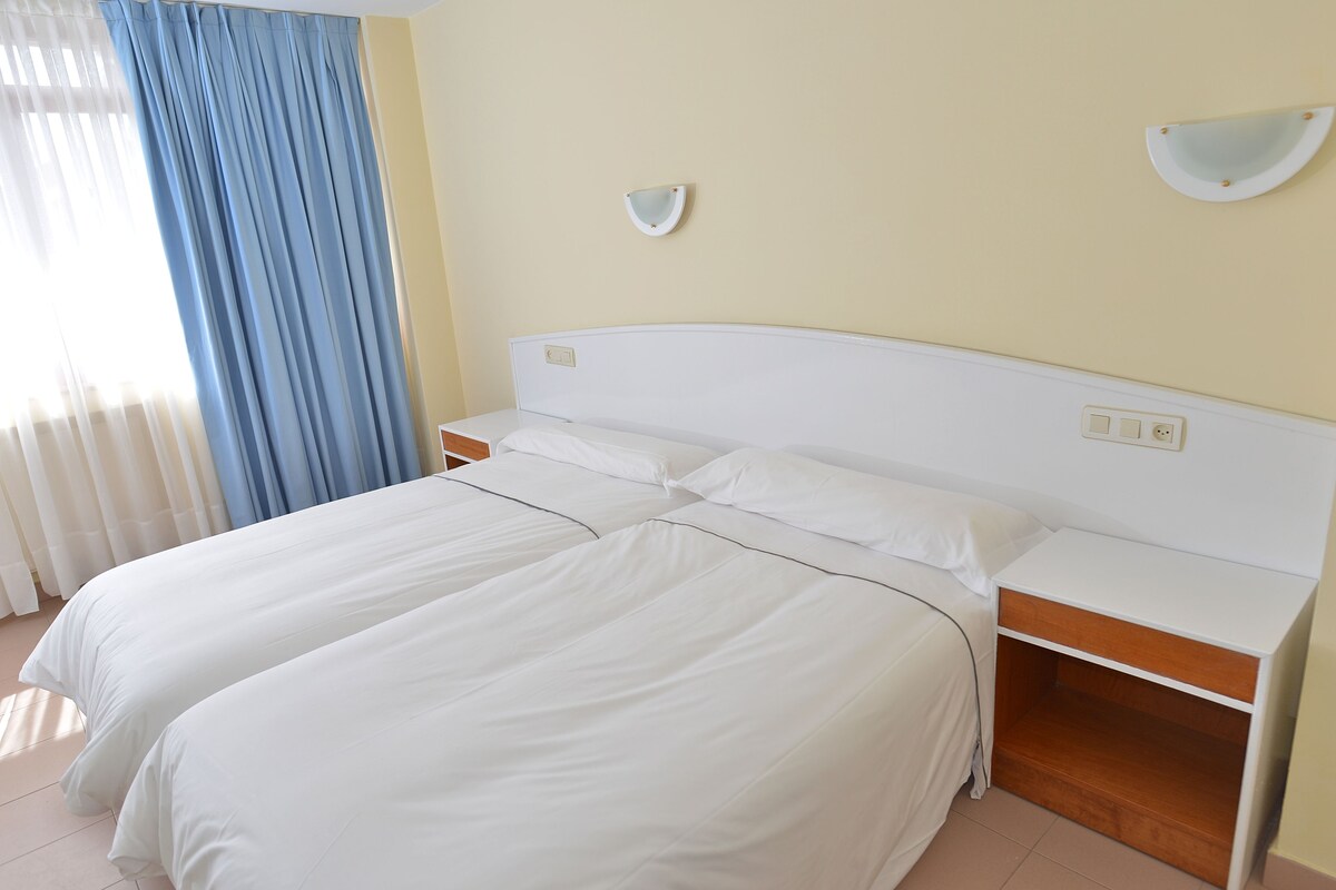 A hotel room with two beds, a nightstand, and a wall-mounted light fixture.