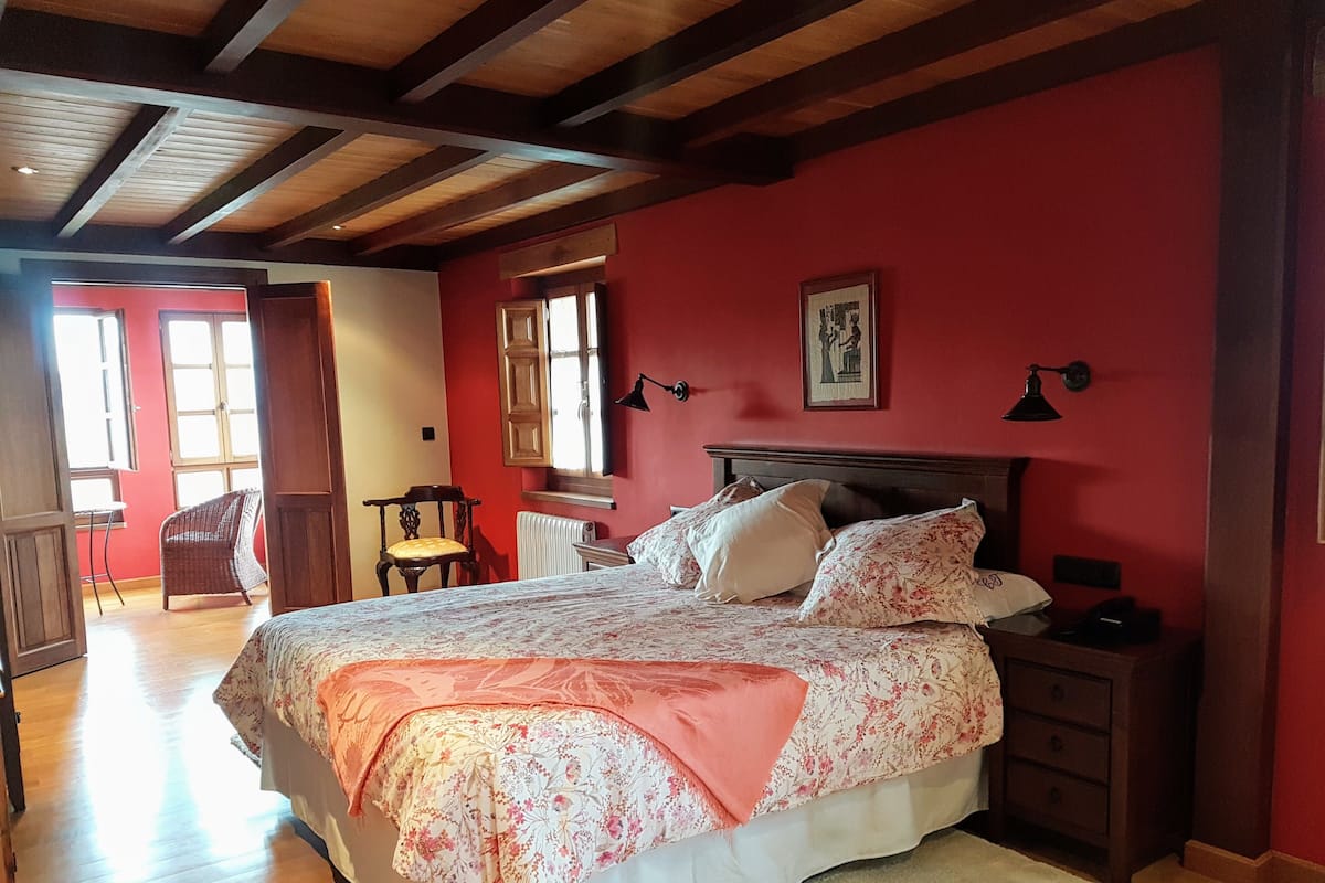 A bedroom with a wooden ceiling, a large bed with floral bedding, a nightstand, and a chair.