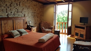 Une chambre avec un mur en pierre, des poutres apparentes en bois au plafond, un lit avec une literie rouge, une chaise, un téléviseur posé sur un meuble en bois et un balcon avec vue.