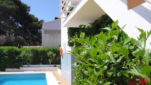 Una piscina con bordo piastrellato, circondata da piante verdi rigogliose e un edificio bianco con balcone piastrellato.