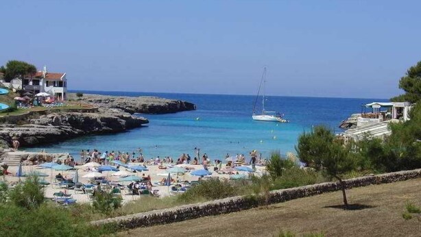Una spiaggia con acque cristalline, un litorale roccioso e una barca all'orizzonte. Si vedono diversi ombrelloni e persone sulla spiaggia.