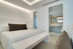 Un dormitorio moderno con una cama blanca, un cojín marrón y una puerta de cristal que da a un baño.