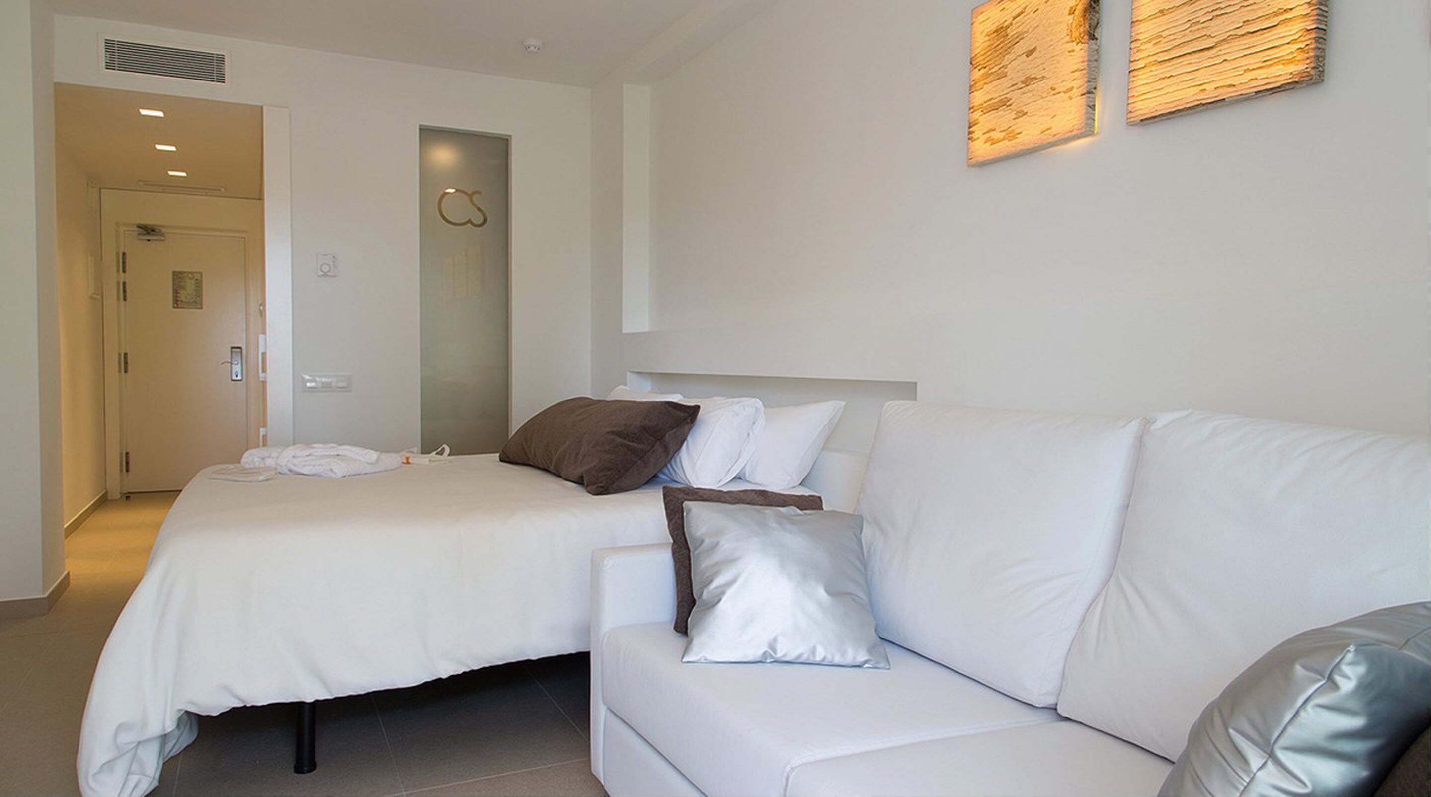 A hotel room with a white bed, a white sofa, and a door leading to a hallway.