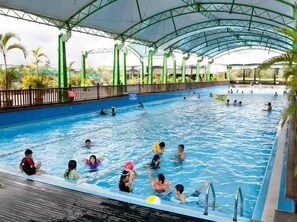 Una piscina interior con techo cubierto, rodeada de palmeras y una terraza de madera.