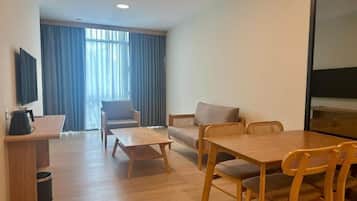 Quarto de hotel com mesa de jantar de madeira, dois sofás, televisão e janela com cortinas.