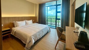 Quarto de hotel com cama, escrivaninha com telefone, televisão e vista para uma varanda com guarda-corpo.