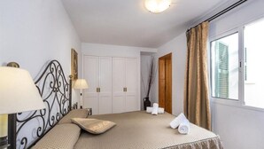 A bedroom with a bed, a nightstand, a lamp, a wardrobe, and a window with curtains. - Villas Geisan (Ciutadella de Menorca)