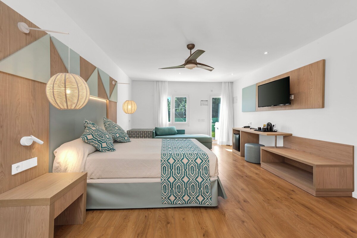 Um quarto moderno com uma cama, mesas de cabeceira, ventoinha no teto, televisão e uma janela com cortinas.