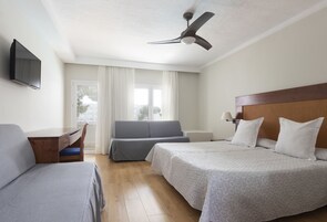 Quarto de hotel com duas camas, uma escrivaninha, uma cadeira, um ventilador de teto e uma televisão.