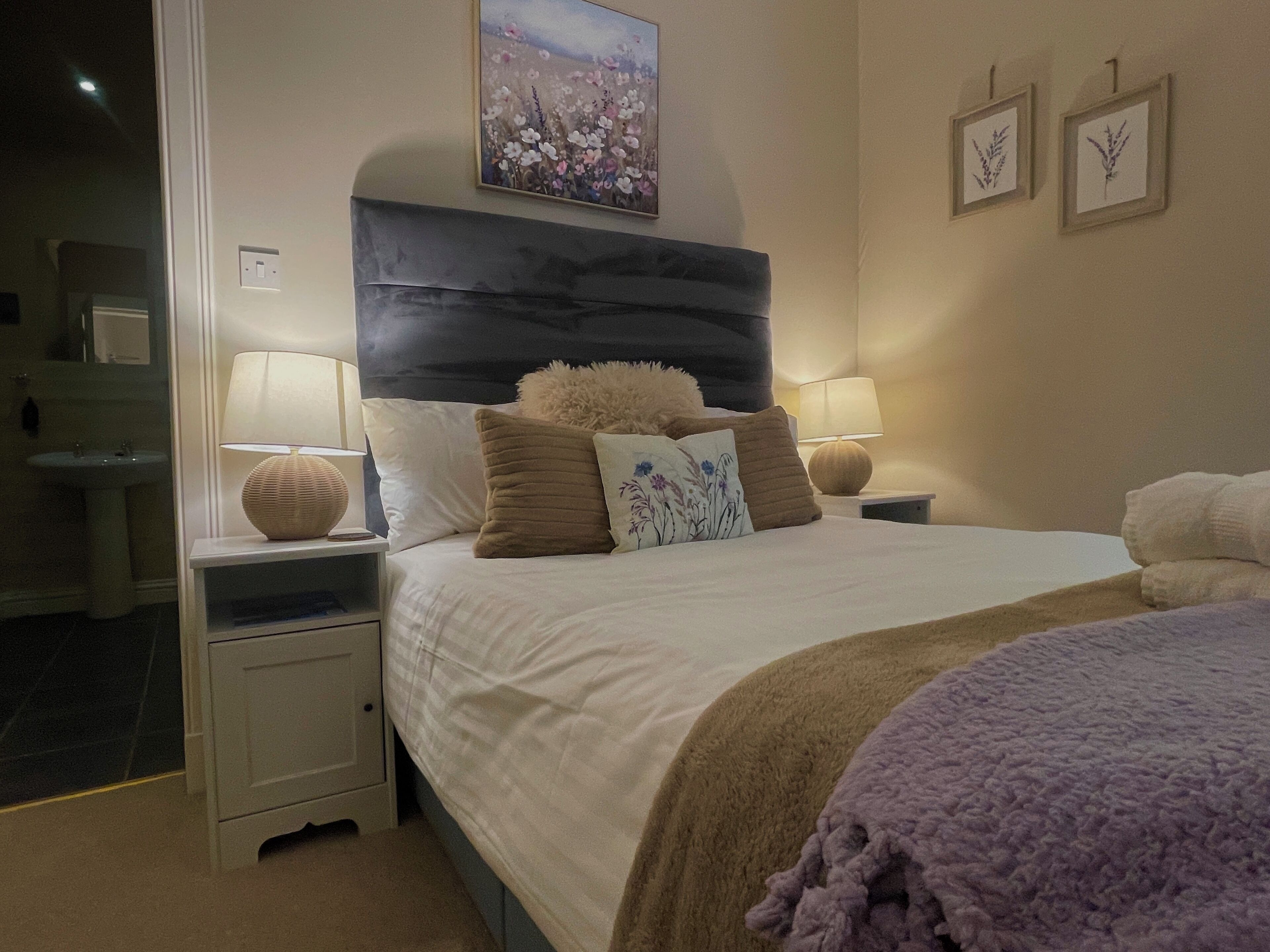 A hotel room with a large bed, two bedside tables with lamps, a nightstand, a wall-mounted painting, and framed botanical prints.