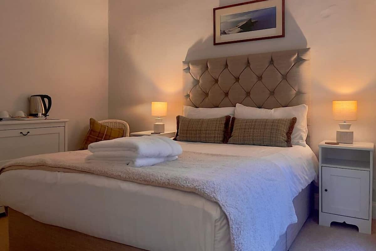 Un lit bien fait, recouvert d’une couette blanche moelleuse, deux lampes de chevet, un tableau encadré au mur et une petite table de chevet avec une bouilloire.