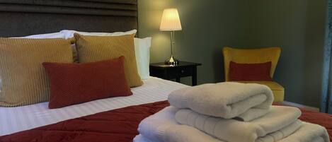 A neatly made bed with a red quilt, pillows in various colors, a lamp on a bedside table, and a chair with a cushion.