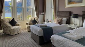 A hotel room with two beds, a sofa, and a chair. There are large windows with curtains, a painting on the wall, and a view of mountains. - Creag Mhor Lodge (Fort William)