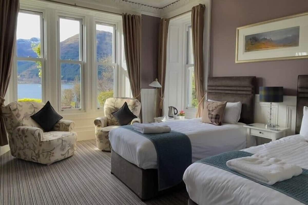 Une chambre d’hôtel avec deux lits, un canapé et un fauteuil. Elle est dotée de grandes fenêtres avec des rideaux, d’un tableau au mur et offre une vue sur les montagnes.