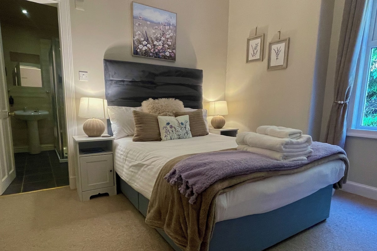 Une chambre à coucher avec un lit, des tables de chevet, des lampes, une commode, une fenêtre avec des rideaux et un tableau au mur.