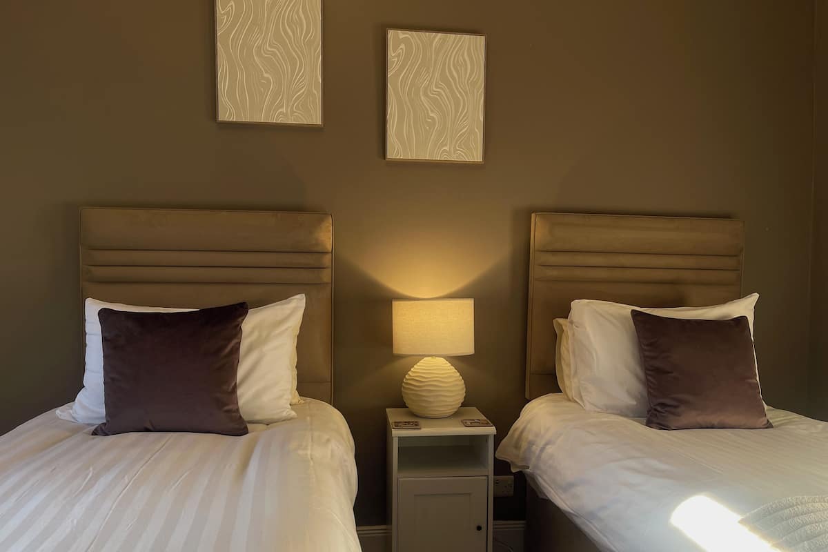 Une chambre d’hôtel avec deux lits, chacun doté d’une tête de lit, d’une table de chevet avec une lampe et d’une petite table de nuit avec un tiroir.