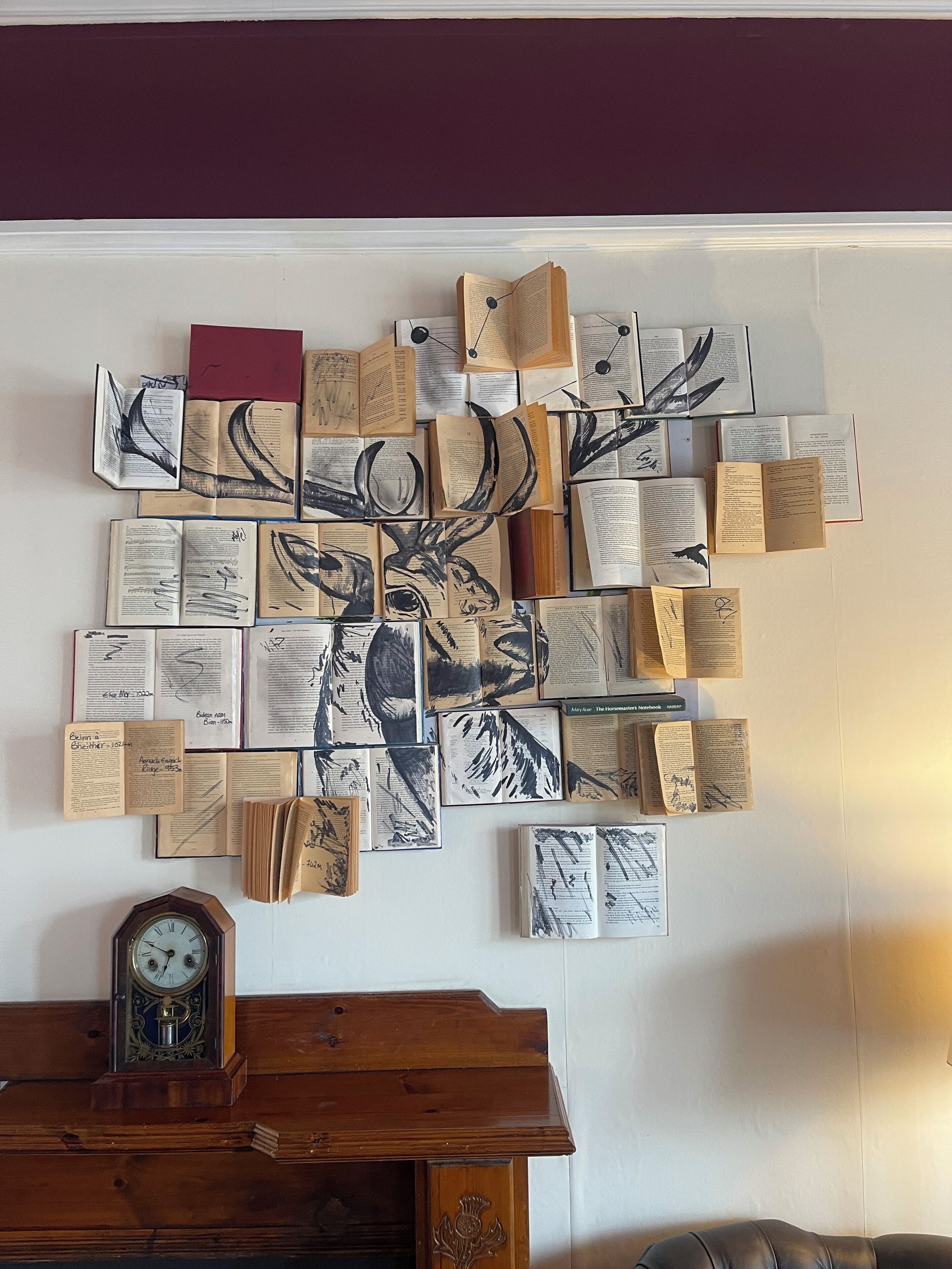 A wall with a large, abstract art piece made of open books and papers. Below the artwork, there is a wooden clock on a wooden cabinet.