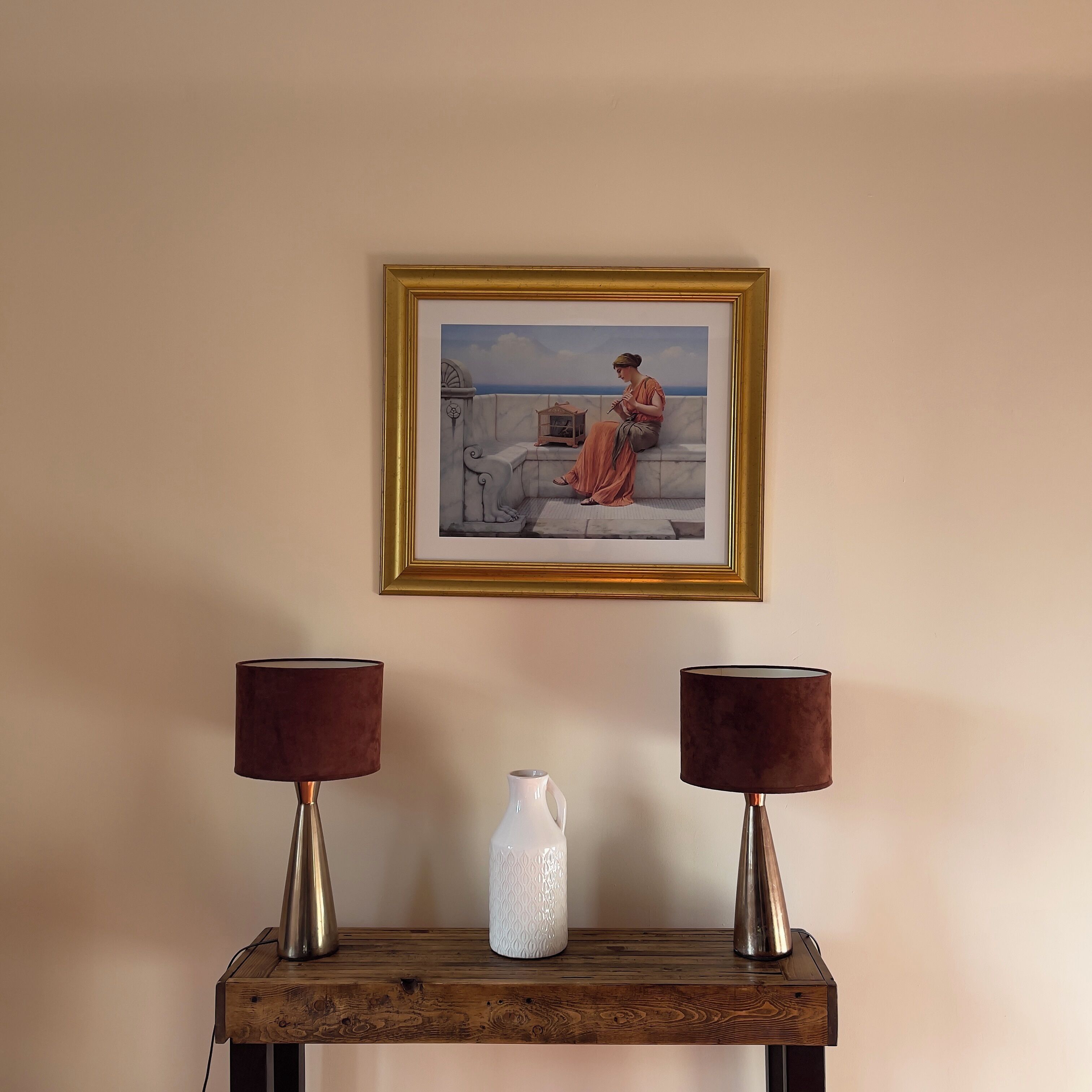 A framed painting of a woman on a balcony, two lamps, and a white vase on a wooden table against a plain wall.