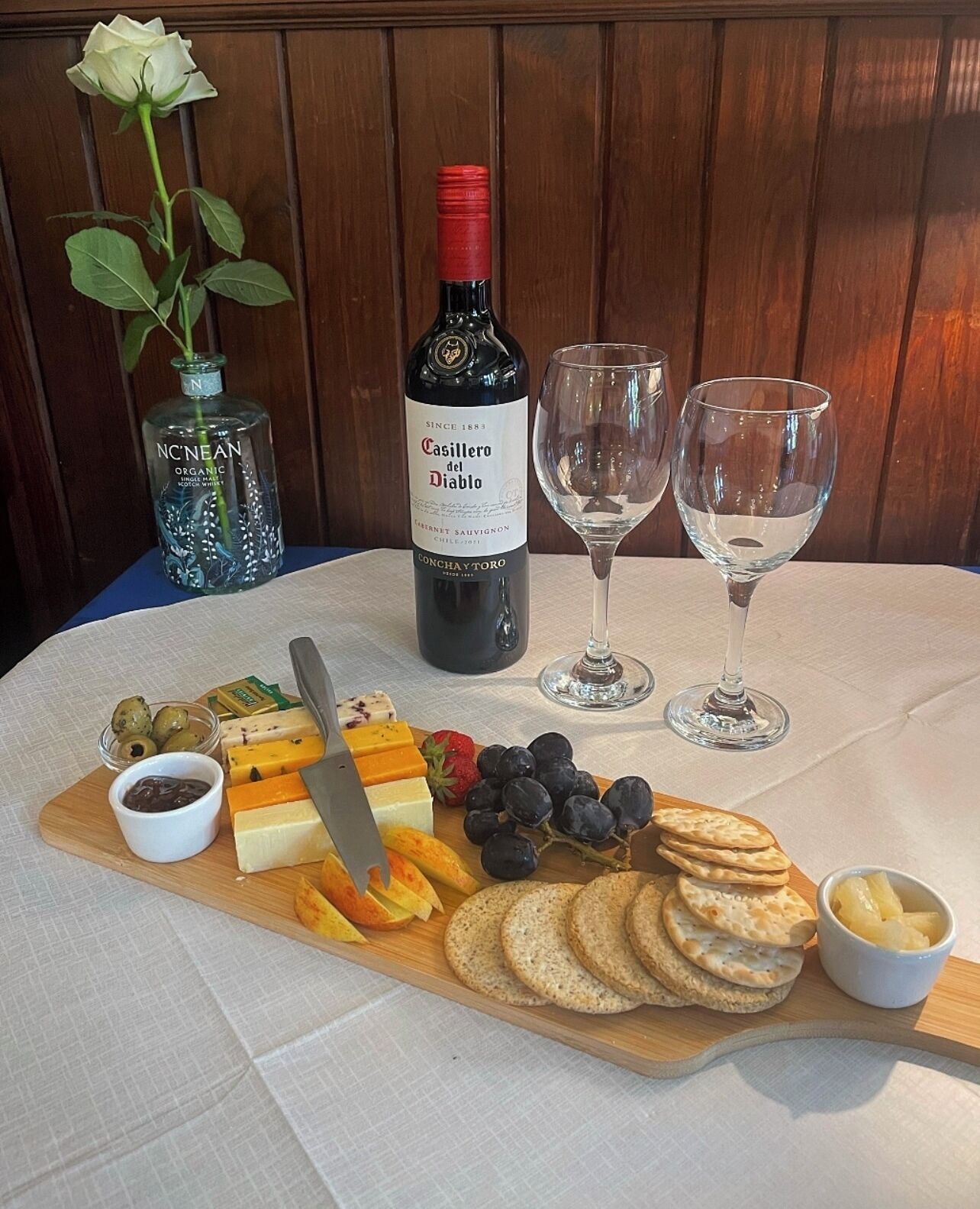 A table with a bottle of Casillero del Diablo wine, two empty wine glasses, a variety of cheeses, grapes, crackers, and olives.