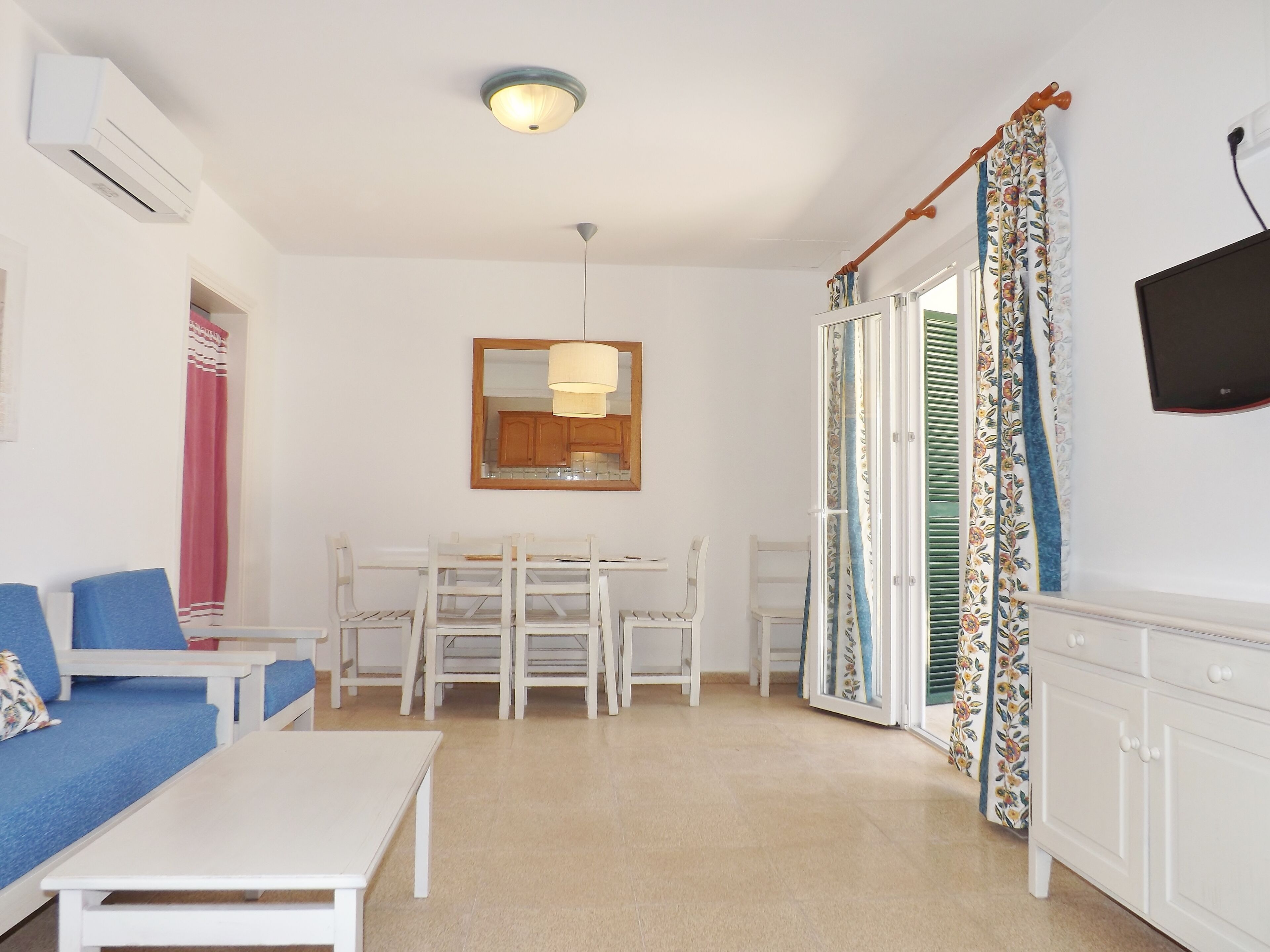 Una habitación con un sofá blanco, una mesita, una mesa de comedor con sillas, una lámpara de techo, un espejo y un televisor en la pared.