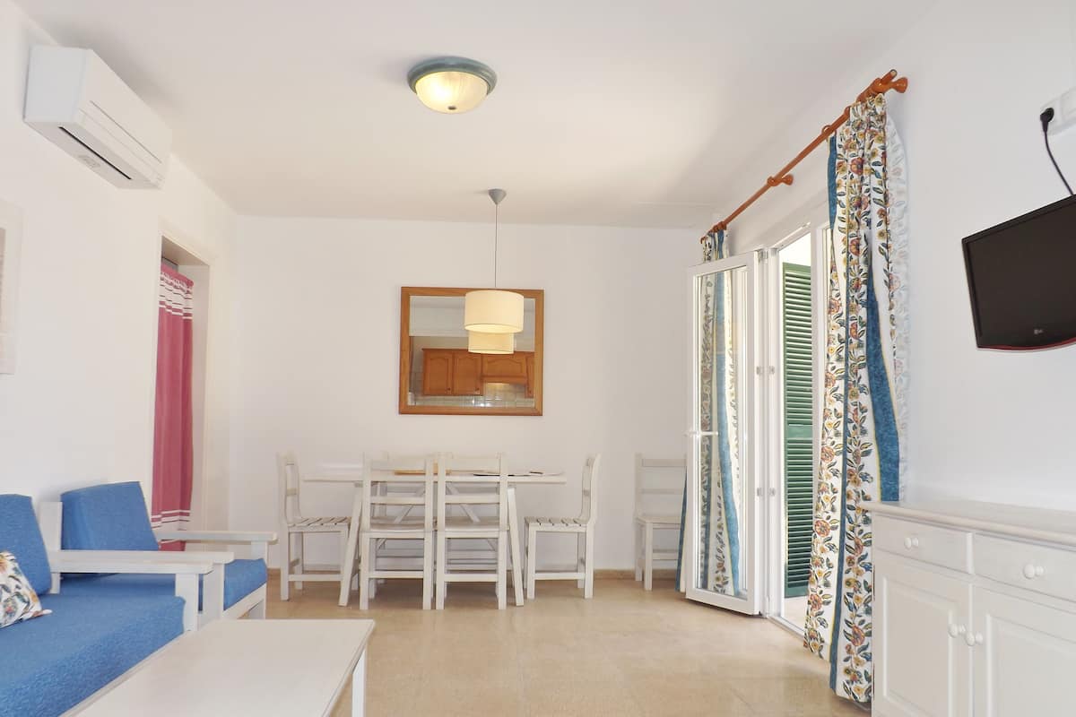 A room with a white sofa, a small table, a dining table with chairs, a ceiling light, a mirror, and a TV on the wall.