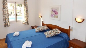 Une chambre d’hôtel avec un lit, deux oreillers, une couvre-lit bleue, deux serviettes enroulées, une fenêtre avec des rideaux, une lampe fixée au mur et un tableau encadré.