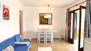 Un salon avec un canapé bleu, une table basse blanche, une table à manger avec des chaises, un miroir et un coin cuisine comprenant un meuble et une fenêtre avec des volets.
