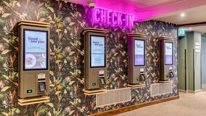 A check-in area with three self-service kiosks against a floral wallpaper backdrop, a neon 'CHECK-IN' sign, and a visible exit door. - Premier Inn Glasgow Stepps - M80 J3 (Glasgow)
