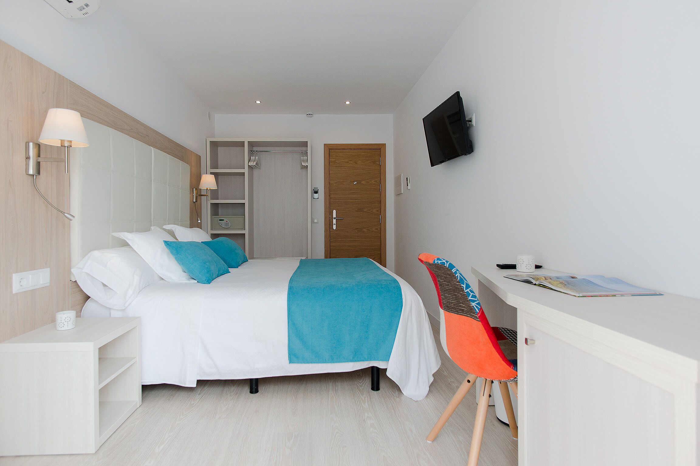 A modern hotel room with a bed, a desk, a chair, and a TV on the wall.