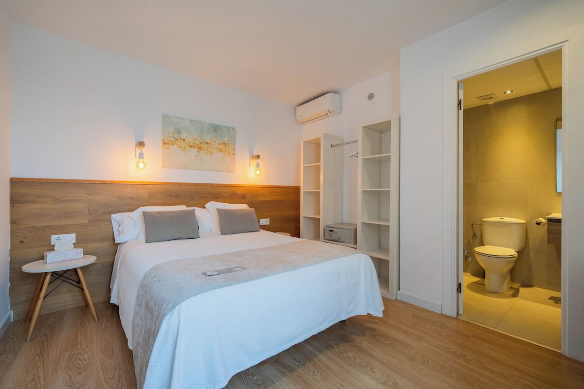 Une chambre d’hôtel avec une tête de lit en bois, un lit avec une literie blanche, une petite table de chevet avec une boîte de mouchoirs, un climatiseur fixé au mur et une salle de bain avec des toilettes et un lavabo.