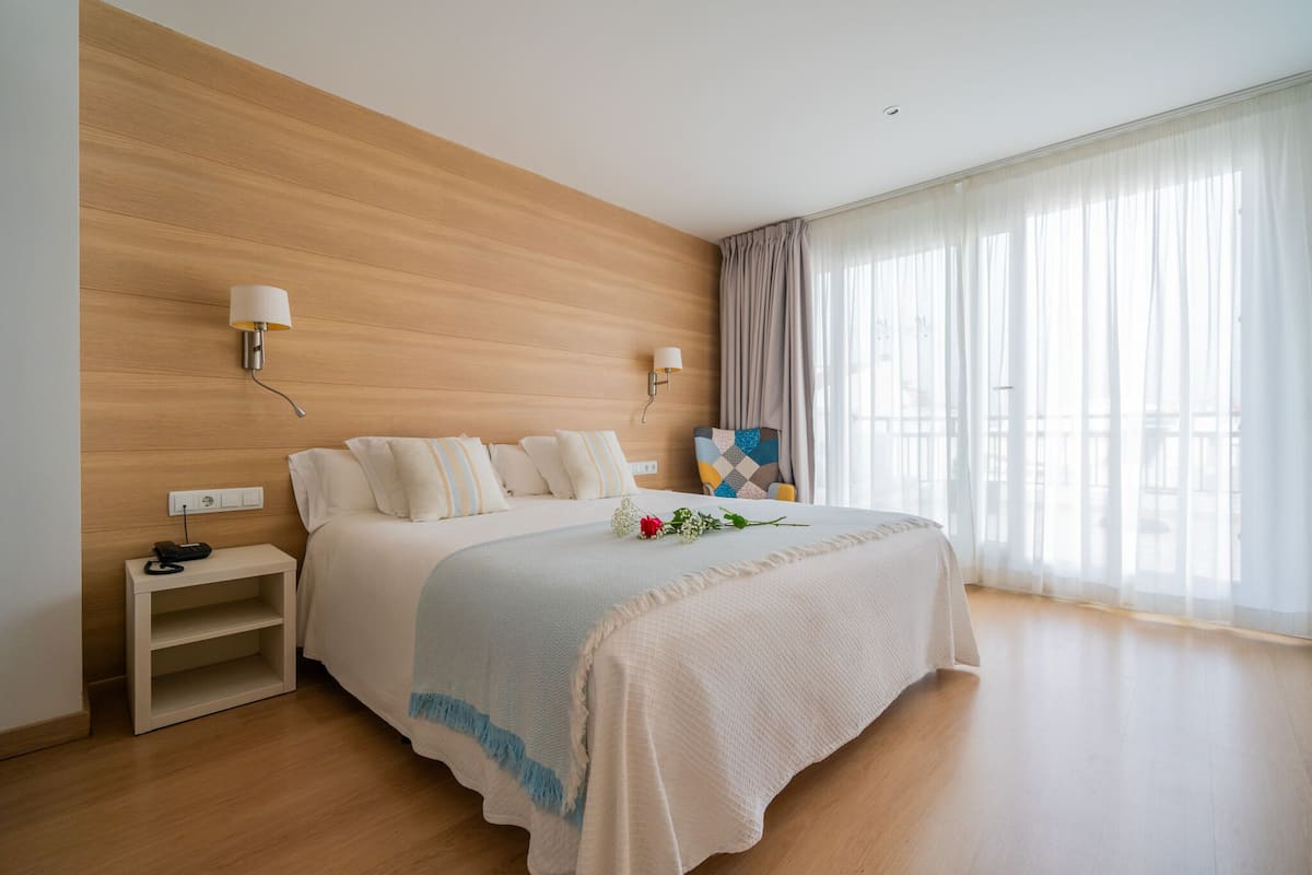 Une chambre d’hôtel moderne avec une tête de lit en bois, un grand lit avec une literie blanche, deux lampes fixées au mur, une table de chevet et une vue sur la ville à travers des rideaux transparents.