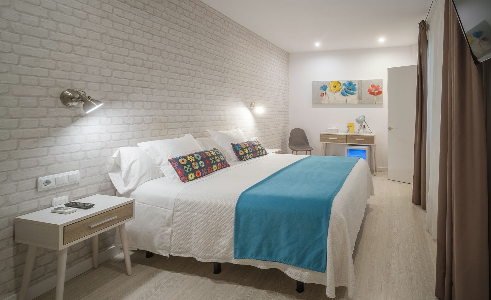 Un dormitorio con pared de ladrillo blanco, cama con ropa de cama blanca y almohadas de colores, un escritorio con lámpara y un cuadro en la pared.