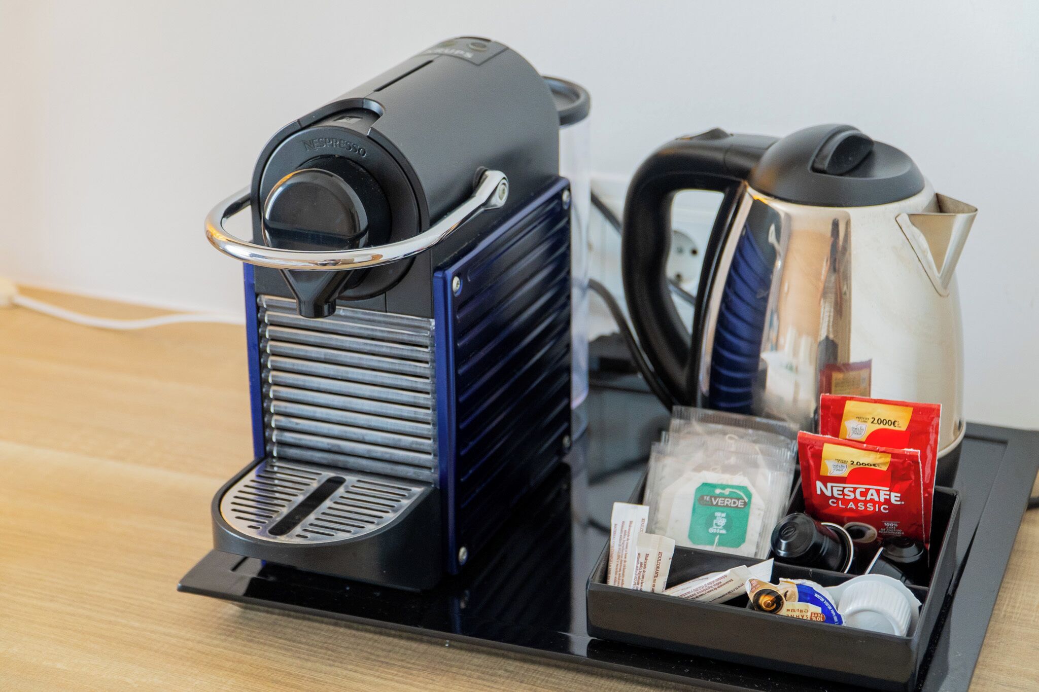 Una cafetera con dispensador de cápsulas Nespresso, una tetera y una bandeja con cápsulas de café y un paquete de café Nespresso Classic.