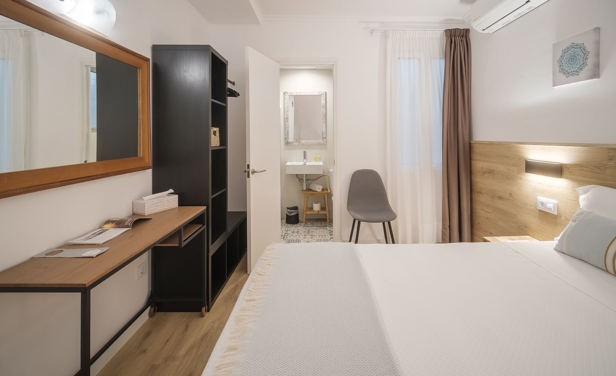 Hotellihuone, jossa on sänky, työpöytä, vaatekaappi, peili, tuoli, kylpyhuone ja ikkuna, jossa on verhot.