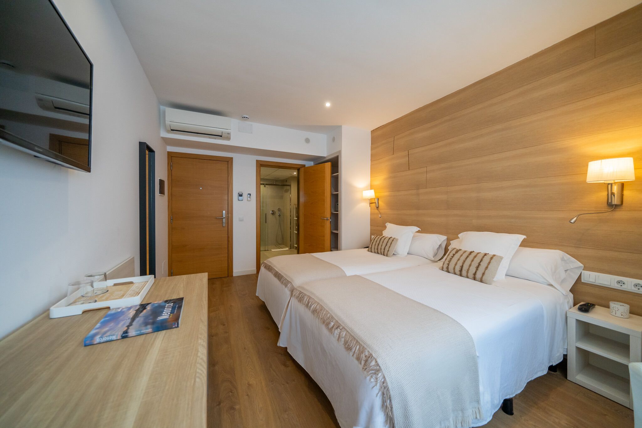 Habitación de hotel con dos camas, cabecero de madera, televisor, mesita de noche con lámpara y zona de ducha.