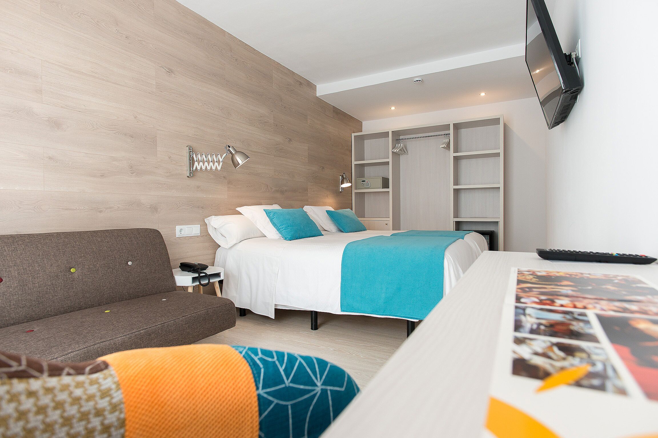 Ein modernes Hotelzimmer mit einer Wand in Holzoptik, einem Bett mit einer türkisblauen Decke, einer Couch, einem an der Wand montierten Fernseher und einem Schreibtisch mit einer Fernbedienung.