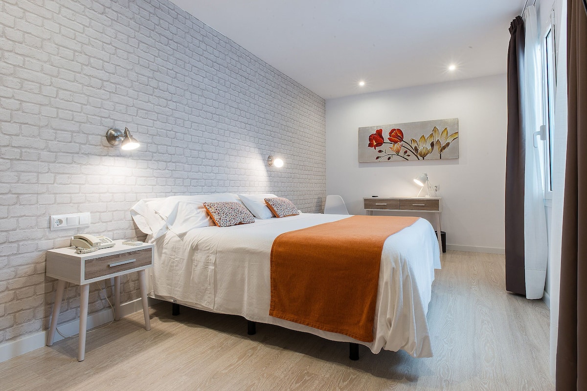 Une chambre moderne avec un mur en briques blanches, un lit avec une literie blanche et un plaid orange, une table de chevet avec une lampe, et un tableau accroché au mur.