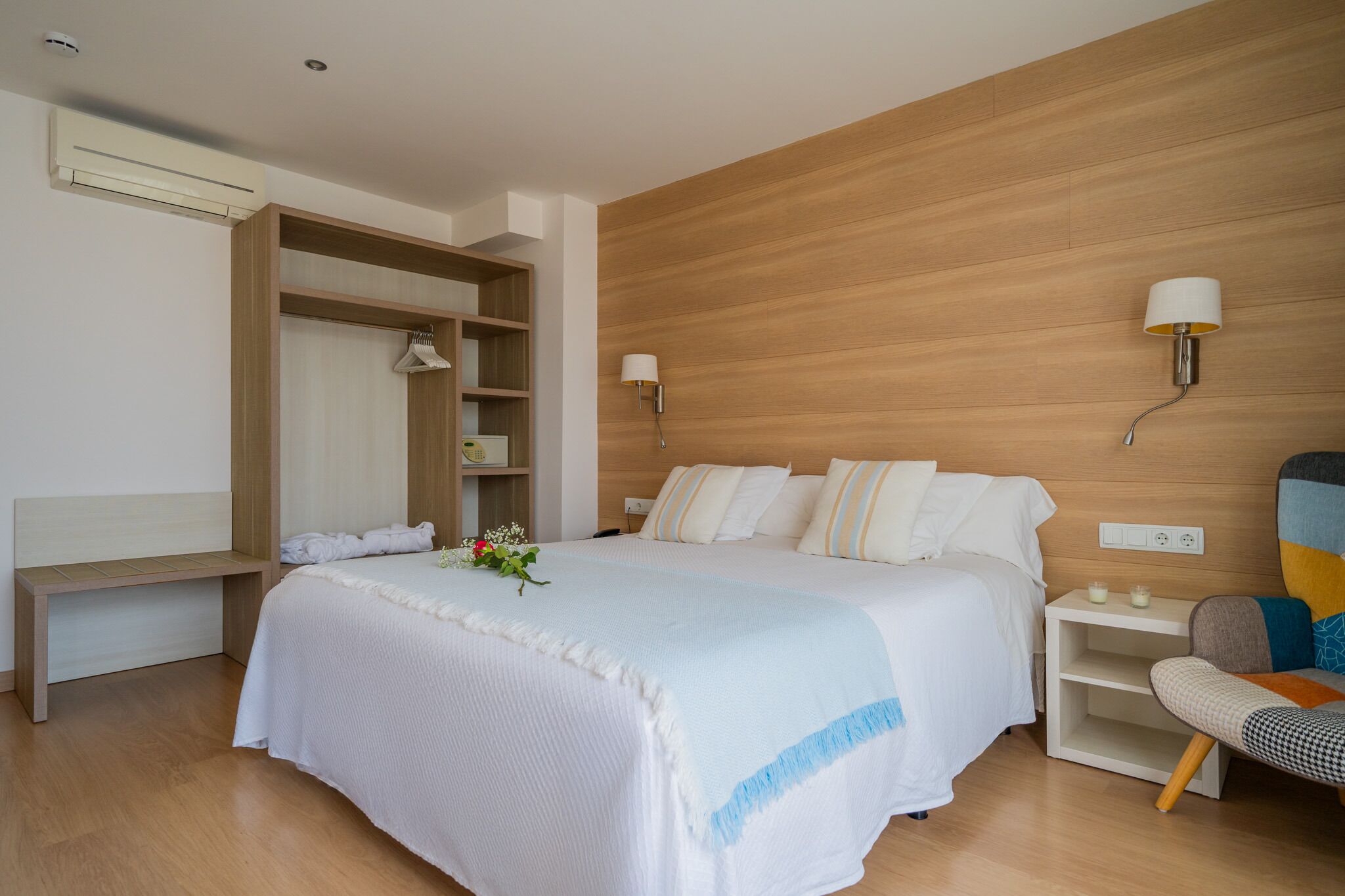 Una habitación de hotel moderna con una cama grande, cabecera de madera, mesita de noche con lámpara, un banco y un armario con espejo.