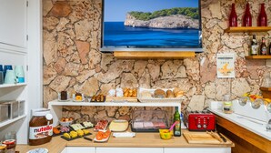 Une cuisine aux murs en pierre, un téléviseur diffusant un paysage marin et diverses denrées alimentaires disposées sur des étagères.