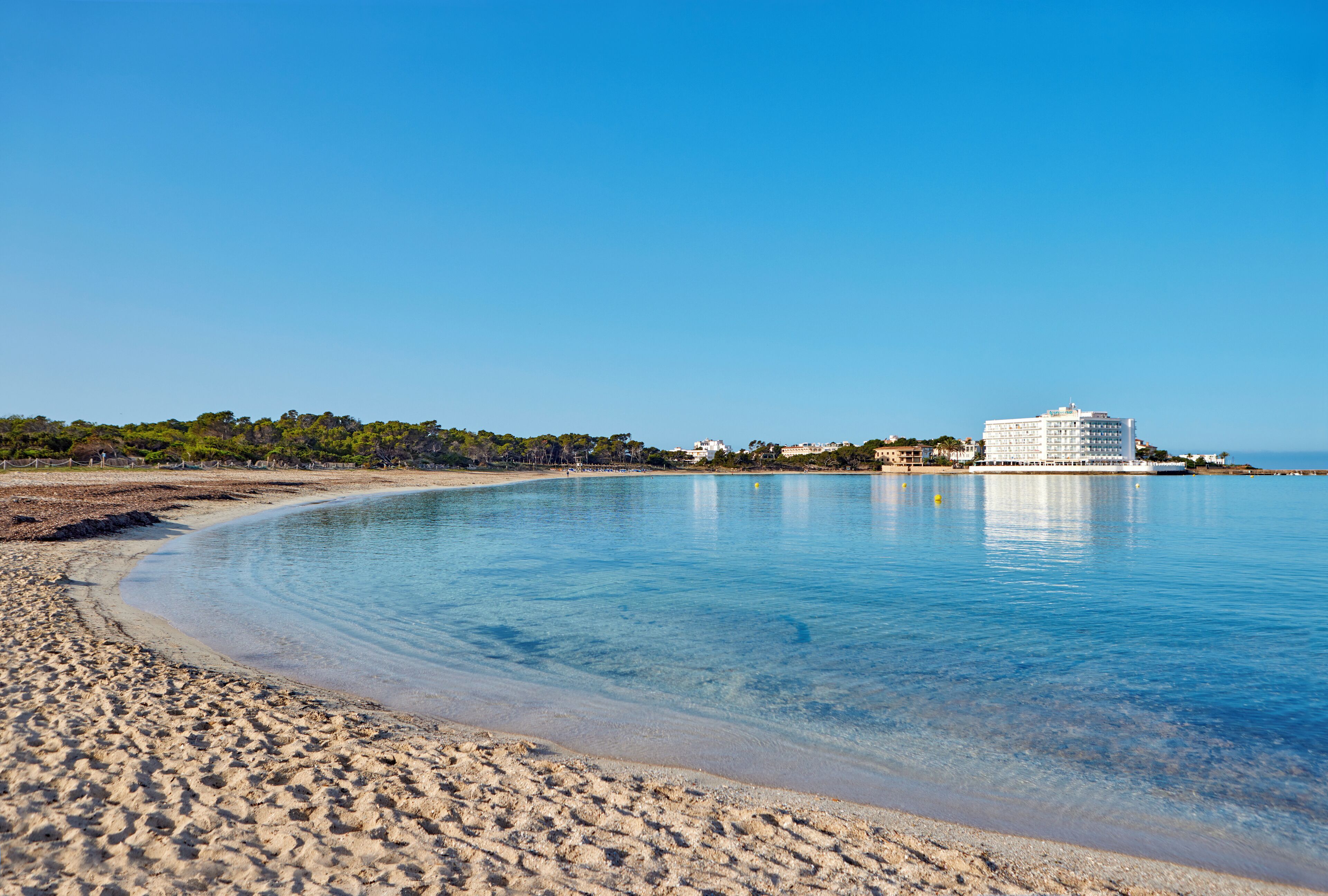 Une plage avec une eau d’un bleu limpide, un rivage sablonneux et un bâtiment blanc en arrière-plan.