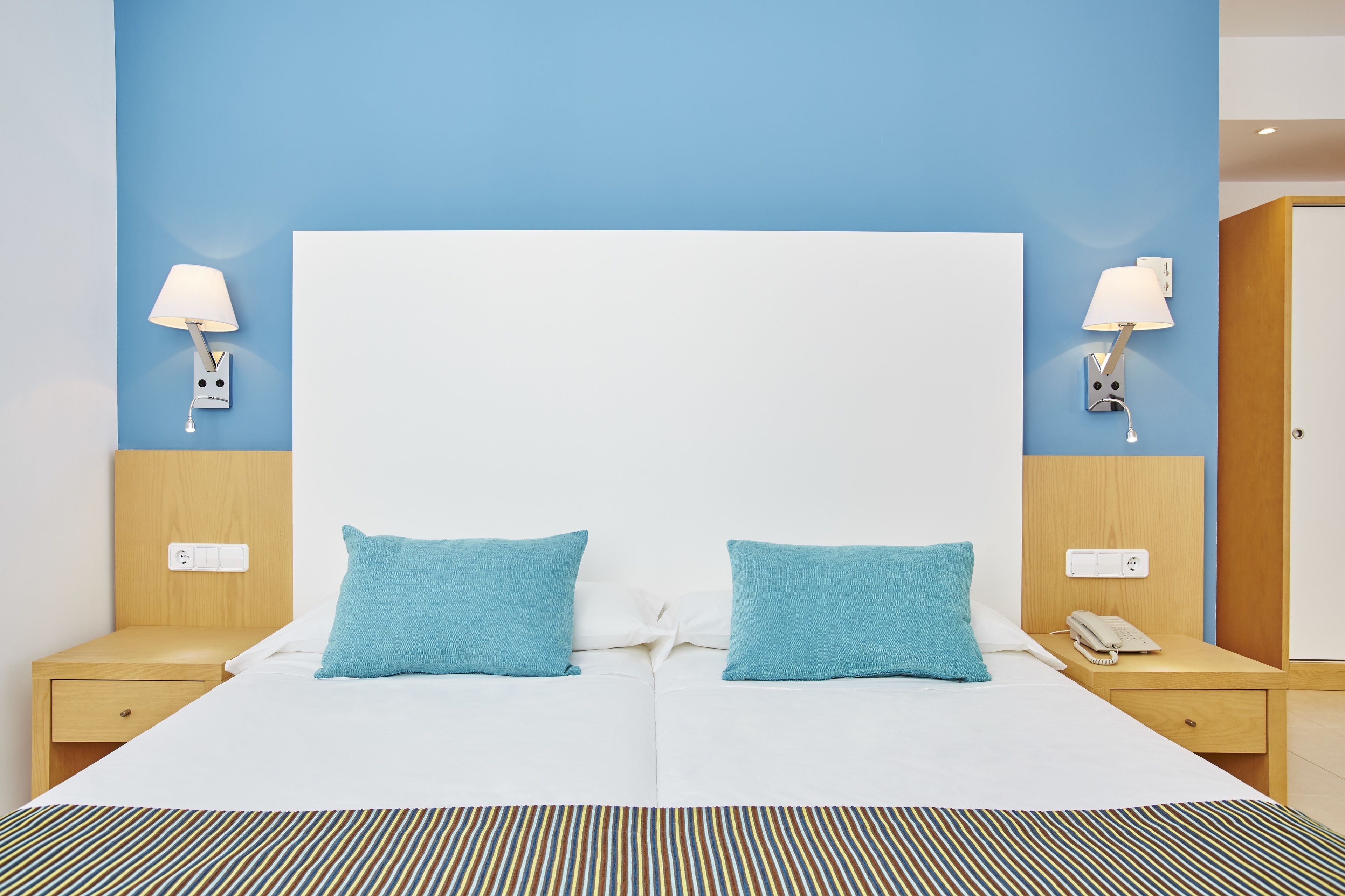 A double bed with two blue pillows, a white duvet, and a striped bedspread. There are two bedside tables with lamps, a phone, and a power outlet on each. The room has a blue accent wall and a wooden headboard.