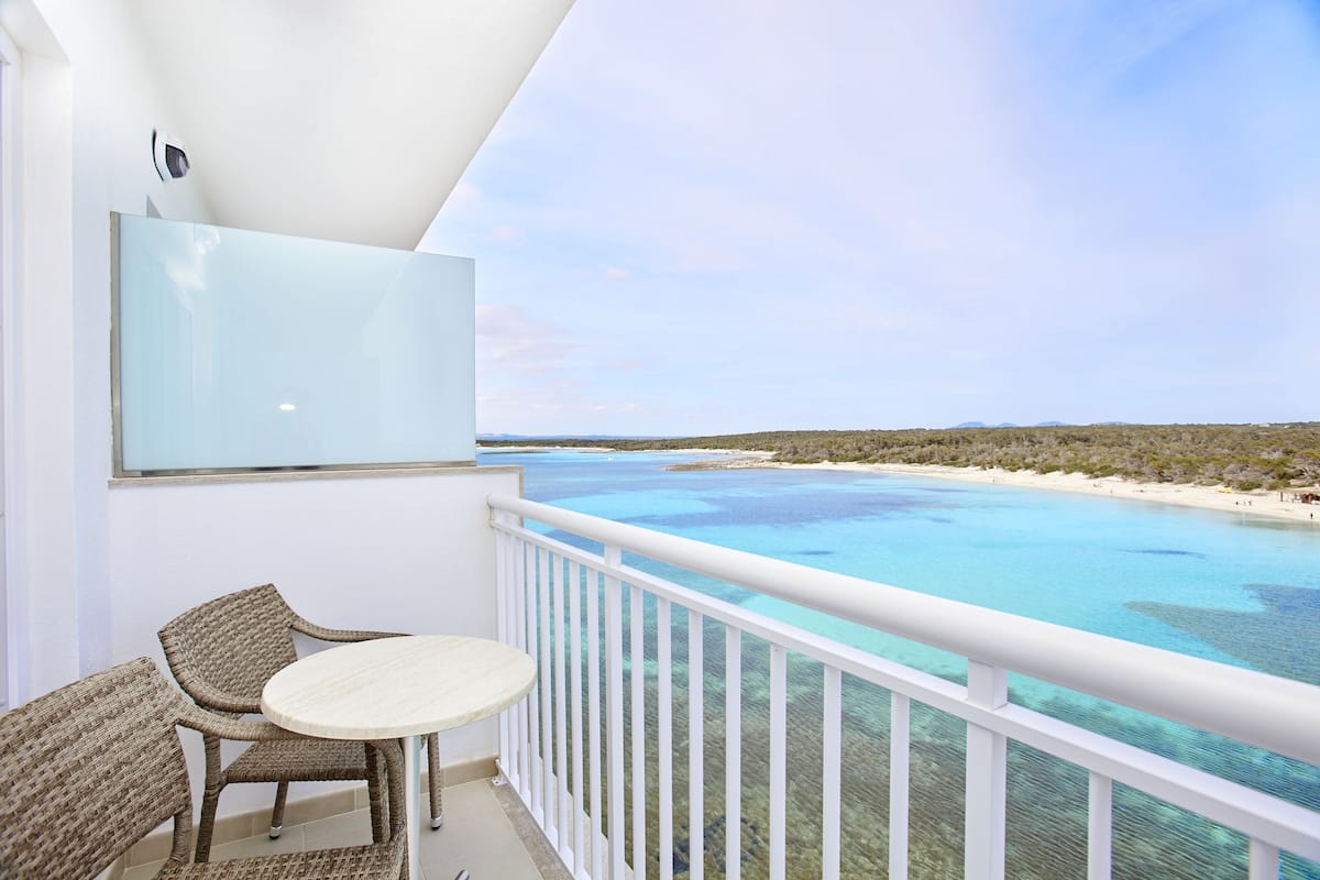 A balcony with wicker chairs and a small table, overlooking a clear blue sea and a sandy beach.