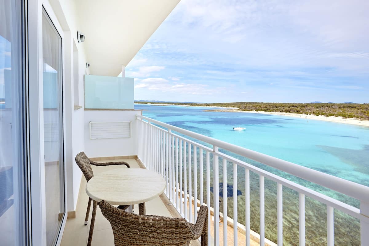 A balcony with a table and chairs, overlooking a clear blue sea and sandy beach.