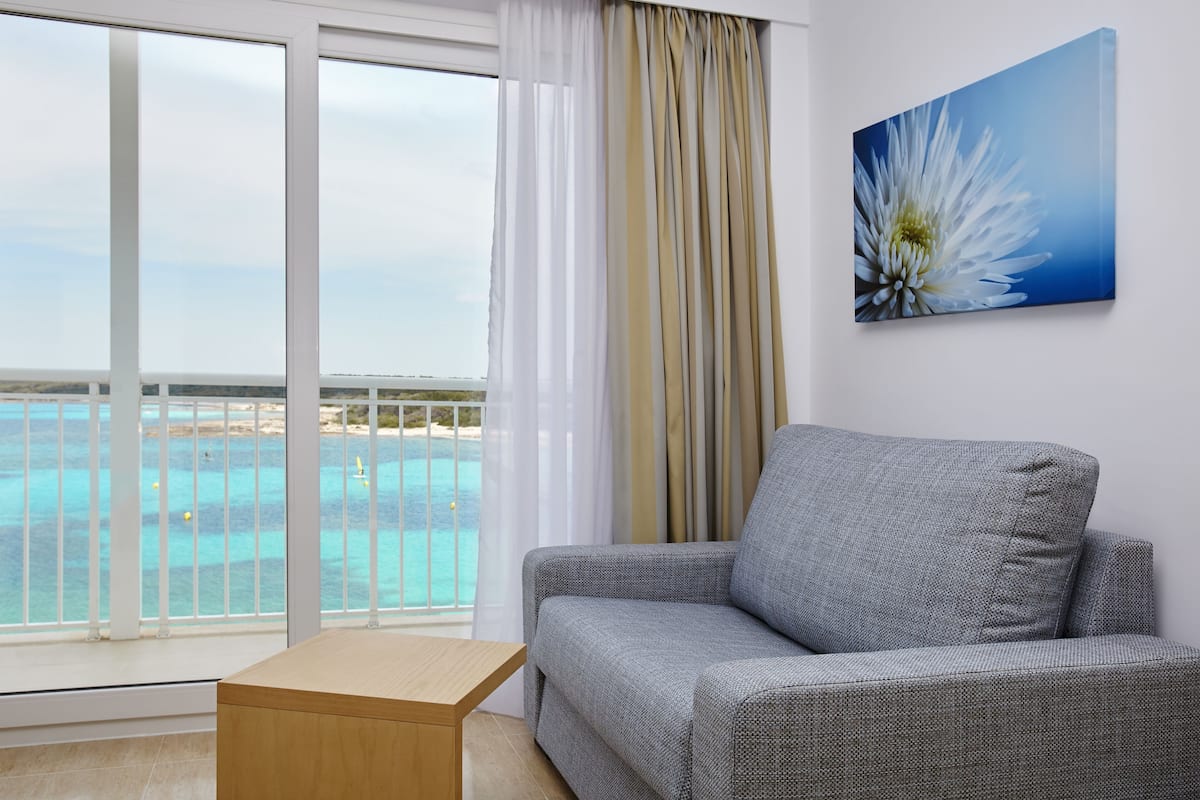 A room with a grey sofa, a wooden side table, a large window with a view of a beach and turquoise waters, and a painting of a white flower on the wall.