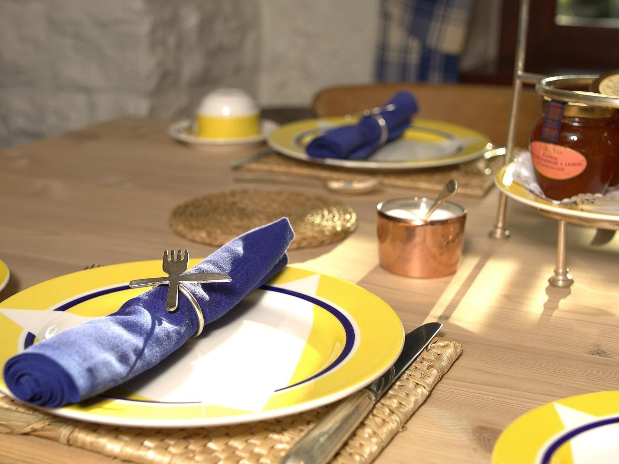 Ein Tisch gedeckt mit gelben und blauen Tellern, einer Gabel, einem Messer, einem Servietttuch, einem kleinen Kupfertopf und einem Marmeladenglas.